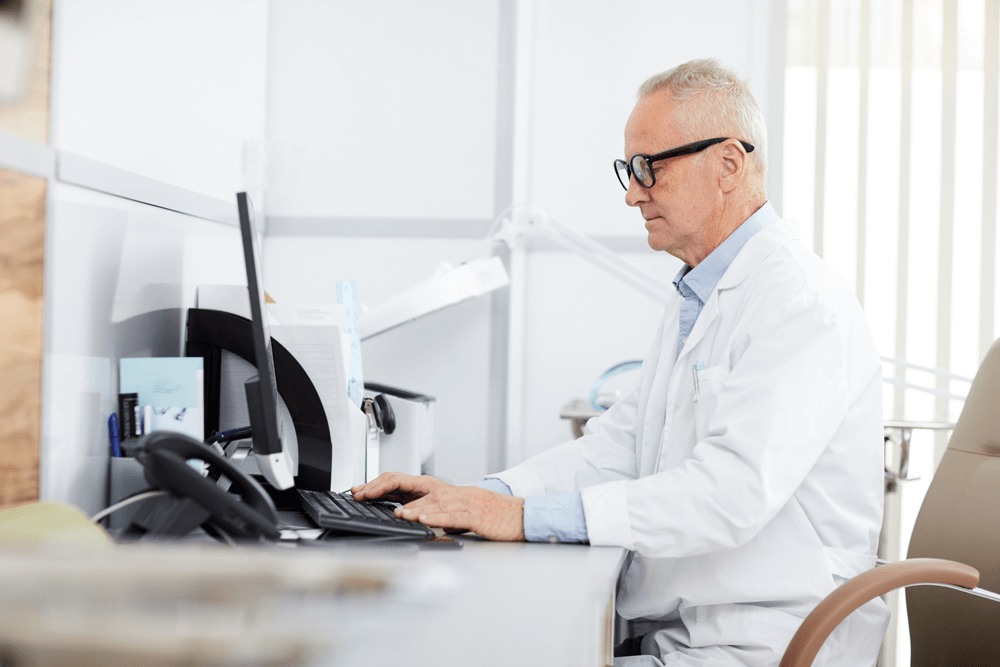A doctor in a white lab coat sits at a desk working on a computer, surrounded by office supplies and equipment.
