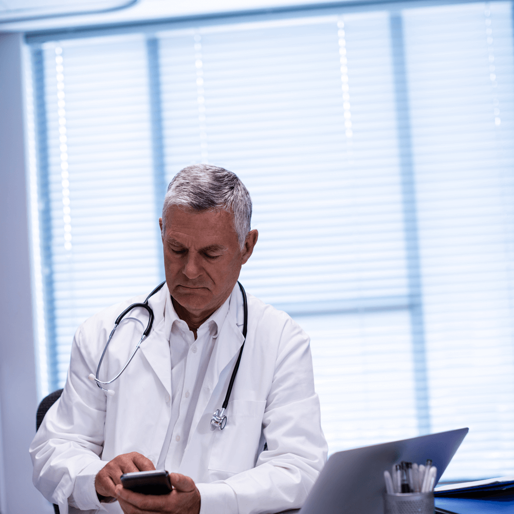 doctor-sitting-at-tablet-and-using-mobile-phone-2025-04-03-15-37-10-utc A doctor in a white coat checks a smartphone while seated at a desk with a laptop and office supplies in a bright, modern office.