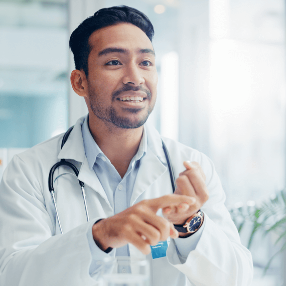 happy-man-doctor-and-consulting-with-patient-for-2025-04-06-10-20-29-utc A doctor in a white coat gestures while discussing in a bright office, with shelves and a plant in the background.