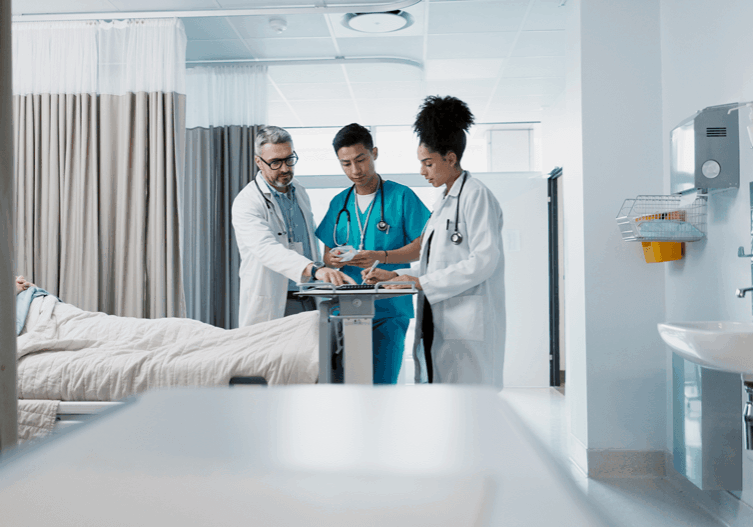 Three medical professionals examine a patient chart near a hospital bed in a bright, sterile healthcare environment.