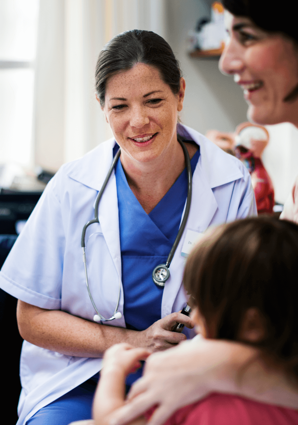 young-patient-getting-checked-by-a-doctor-2025-02-10-11-37-53-utc A healthcare professional in a white coat with a stethoscope talks to a young child while a caregiver supports the child.